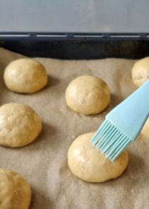 Buns sans gluten au zeste d’orange - Préchauffe le four à 180°C. Dans un petit bol, bats un œuf mélangé avec une cuillerée de lait d’avoine, puis badigeonne les pains avec cette dorure. La dorure aide à obtenir une croûte dorée.
