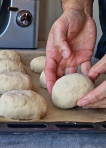 Buns sans gluten au zeste d’orange - Façonne la pâte en formant ta main en « griffe » et en la déplaçant en mouvement circulaire contre la surface. Tire les coins de la pâte et ramène-les vers le milieu pour façonner chaque morceau en boule. Le mouvement circulaire main-surface aidera à former une boule.