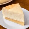 Part de cheesecake new-yorkais sur une assiette blanche, la fine base génoise sous la garniture au fromage frais