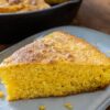 Part de cornbread sur une assiette, la mie de maïs dorée côté coupe, la poêle en fonte derrière